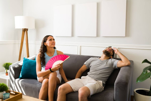 A man and woman with a hand fan sitting on a a couch looking uncomfortable because of the heat inside their home.